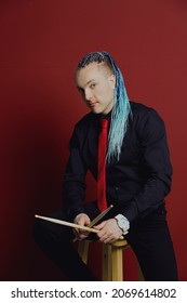 A young attractive man with an interesting hairstyle in pigtails on an undercut with a red tie on a red background in and drumsticks in his hands.