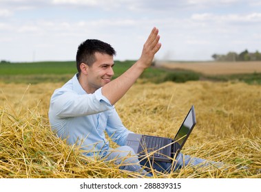 Young attractive man with laptop sitting on haystack at farmland and waving hand to someone