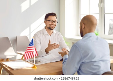A young attractive man sitting in the office of the US public services or embassy with immigration application and having consular visa interview with US flag at the desk of official's workplace. - Shutterstock ID 2286749955