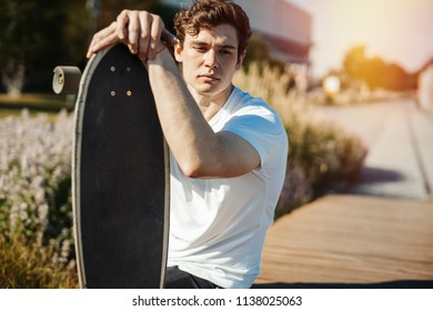 Young attractive man sitting on the bench in the park and holding longboard.