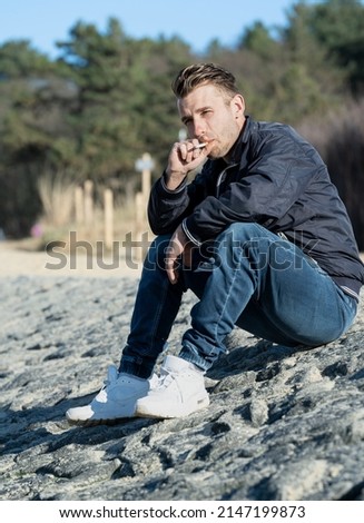 Young attractive man smokes a cigarette. Sitting. Beach and stones in the background.

