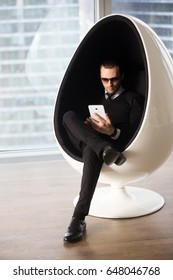 Young attractive man in stylish wear and sunglasses working on tablet computer while sitting in futuristic egg chair. Successful businessman managing company or shopping online, entertains in Internet