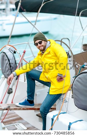 Young attractive man in a yellow waterproof jacket in a hat and glasses plans to go fishing on a boat