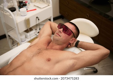 Young attractive manly sporty man wearing UV protective goggles, lying on massage table of spa salon during laser hair removal procedure. Masculine beauty spa treatment , male waxing concept - Shutterstock ID 2073475283