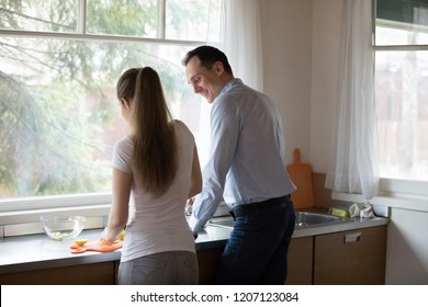 Young Attractive Millennial Couple Standing In The Kitchen Near Countertop At Modern Home Opposite Window, Rear View. Positive Husband And Wife Feels Good Talking And Preparing Cooking Dinner Together