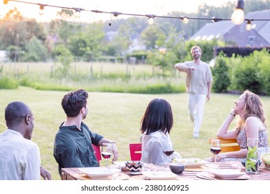 A young, attractive millennial man arrives at a garden dinner party or BBQ to find a multi-ethnic group of friends already enjoying wine and food around the table. As he approaches, the gathering