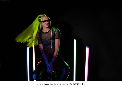 Young Attractive Millennial Woman With Yellow Hair Wearing Modern Glasses Sitting Near Colorful Neon Tube Lights And Posing On Camera. Black Background. Fashionable Futuristic Neon Portrait. 