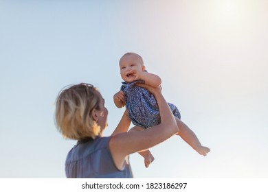 Young attractive mother dandling her happy baby girl while hanging out outdoors. Young woman holding a baby girl on hands against blue sky - Shutterstock ID 1823186297