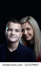 A Young Attractive Mother Is Standing Behind Her White Teenage Son And Both Are Smiling. Photo On A Dark Background