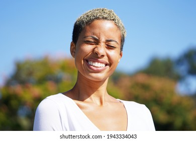 Young Attractive Multicultural Woman Outside In Balboa Park Against The Sky And Trees With Short Hair And A V Neck White Shirt In The Sun, Wincing And Smiling With Eyes Closed  