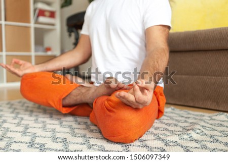Young attractive nice man do yoga practice at home near grey sofa on carpet. Sitting relax meditative asana near couch. Basic yoga workout that  Male wear white t-shirt and orange trouses. Close up
