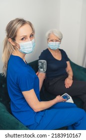 A Young, Attractive Nurse With Blue Eyes And Long Blond Hair, In A Dark Blue Uniform, Measuring The Arterial Blood Pressure Of An Elderly Woman With A Face Mask On During A Pandemic. High-quality