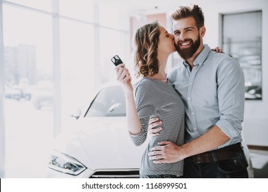 Young Attractive Owners of New Car Hugging. Beautiful Woman Kissing Caucasian Handsome Bearded Smiling Happy Man and Wearing Casual Clothes Standing near Owned Automobile.