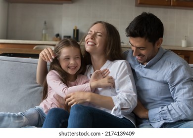 Young attractive parents and preschool adorable daughter play seated on couch, laughing, feeling overjoyed, tickling each other enjoy playtime and carefree active family leisure having fun at home