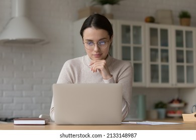 Young attractive pensive woman wear glasses using laptop seated in kitchen, makes remote order, working from home use internet, looking consecrated and thoughtful. Telecommute job, modern tech concept