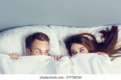 Young attractive playfull couple peeping from bedsheet on the bed at bedroom. Caucasian models in love, relationship, dating, happy people, bedtime concept shot.