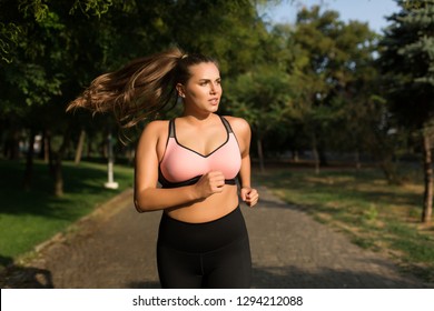Young attractive plus size woman in sporty top and leggings thoughtfully looking aside running in cozy city park 