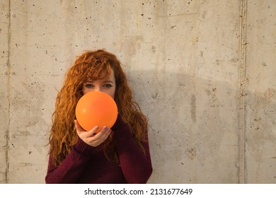 Young, attractive, red-haired, freckled, young woman in red sweater, filling an orange balloon on a gray wall. Concept colors, balloons, beauty, fashion, red.