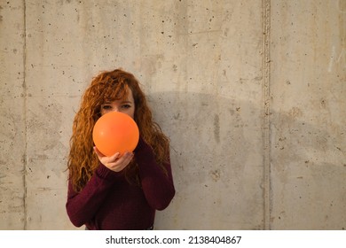 .Young, attractive, red-haired, freckled, young woman in red sweater, filling an orange balloon on a gray wall. Concept colors, balloons, beauty, fashion, red.