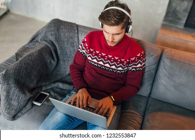 Young Attractive Serious Concentrated Man Sitting On Sofa At Home In Winter, Wearing Red Knitted Sweater, Working On Laptop, Freelancer, Busy, Listening To Headphones, Typing, Studying Online