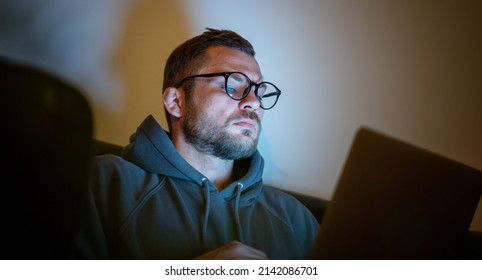 Young attractive serious man in glasses sitting at home at night on an armchair looking into a laptop. Workaholic, insomnia or hacker concept - Shutterstock ID 2142086701