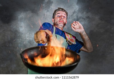 Young Attractive And Shocked Messy Home Cook Man With Apron Holding Pan In Fire Burning The Food In Kitchen Disaster And Unskilled And Unexperienced Terrible Home Cook At Domestic Cooking