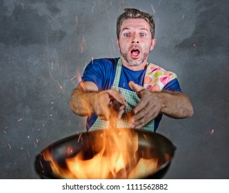 Young Attractive And Shocked Messy Home Cook Man With Apron Holding Pan In Fire Burning The Food In Kitchen Disaster And Unskilled And Unexperienced Terrible Home Cook At Domestic Cooking