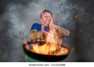 Young Attractive And Shocked Messy Home Cook Man With Apron Holding Pan In Fire Burning The Food In Kitchen Disaster And Unskilled And Unexperienced Terrible Home Cook At Domestic Cooking