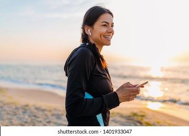 young attractive slim woman doing sport exercises on morning sunrise beach in sports wear, healthy lifestyle, listening to music on wireless earphones holding smartphone, smiling happy