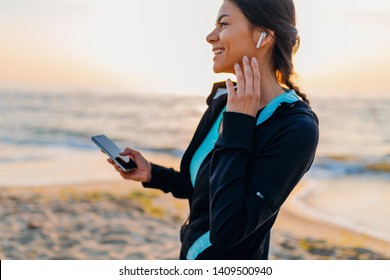 young attractive slim woman doing sport exercises on morning sunrise beach in sports wear, healthy lifestyle, listening to music on wireless earphones holding smartphone, smiling happy having fun