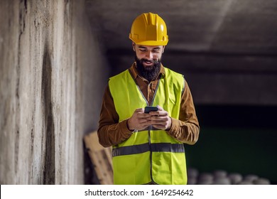 Young attractive smiling bearded worker in vest with helmet on head standing inside of building in construction process, typing a message on smart phone and taking a break from work.