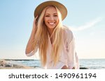 Young attractive smiling blond woman in white shirt and hat joyfully looking in camera with sea on background