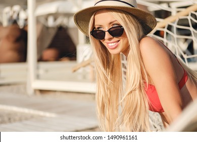 Young attractive smiling blond woman in red swimsuit wearing sunglasses and hat happily looking aside on beach