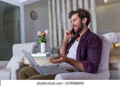 Young Attractive Smiling Guy Is Browsing At His Laptop And Talking Oh Mobile Phone, Sitting At Home On The Cozy Sofa, Wearing Casual Outfit. Freelance Business Work From Home Concept