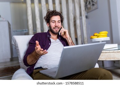 Young Attractive Smiling Guy Is Browsing At His Laptop And Talking Oh Mobile Phone, Sitting At Home On The Cozy Sofa, Wearing Casual Outfit. Freelance Business Work From Home Concept