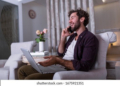 Young Attractive Smiling Guy Is Browsing At His Laptop And Talking Oh Mobile Phone, Sitting At Home On The Cozy Sofa, Wearing Casual Outfit. Freelance Business Work From Home Concept