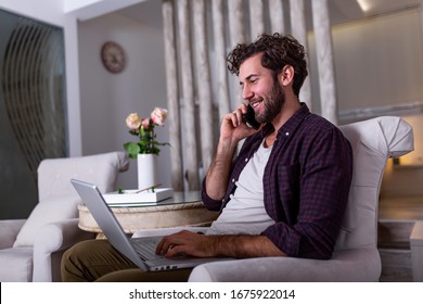 Young Attractive Smiling Guy Is Browsing At His Laptop And Talking Oh Mobile Phone, Sitting At Home On The Cozy Sofa, Wearing Casual Outfit. Freelance Business Work From Home Concept