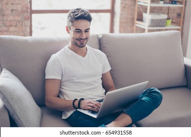 Young attractive smiling guy is browsing at his laptop, sitting at home on the cozy beige sofa at home, wearing casual outfit