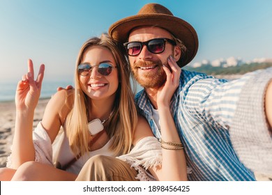 young attractive smiling happy man and woman in sunglasses sitting on sand beach taking selfie photo on phone camera, romantic couple by the sea on sunset, boho hipster style outfit - Powered by Shutterstock