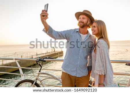 young attractive smiling happy man and woman traveling on bicycles taking selfie photo on phone camera, romantic couple by the sea on sunset, boho hipster style outfit, friends having fun together