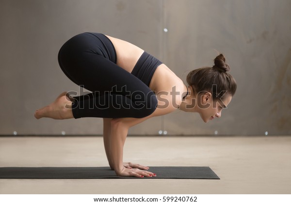 Young attractive smiling yogi woman practicing yoga,\
standing in Bakasana exercise, Crane pose, working out, wearing\
black sportswear, cool urban style, full length, grey studio\
background, side view 