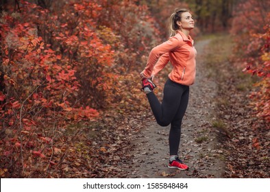 Young attractive sport woman in sportswear doing stretching leg in autumn forest with reddened leaves of trees. Warm up before running. Healthy lifestyle concept