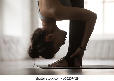 Young attractive sporty woman practicing yoga, standing forward bend exercise, head to knees, uttanasana pose, working out, wearing sportswear, white loft studio background, side view, close up - Powered by Shutterstock