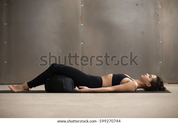 Young attractive sporty yogi woman practicing yoga,\
lying in Savasana exercise, Dead Body, Corpse pose using Zafu\
cushion for comfort, resting after working out, urban style grey\
studio, full length 