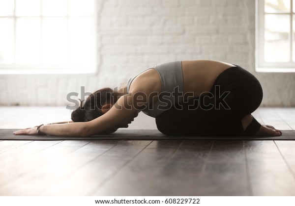 Young attractive sporty yogi woman practicing yoga\
concept, sitting in Child exercise, Balasana pose, working out,\
wearing black sportswear, full length, white loft studio\
background, side view