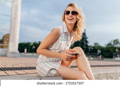 Young Attractive Stylish Blonde Woman Sitting In City Street In Summer Fashion Style Dress Wearing Sunglasses, Holding Phone, Laughing Candid