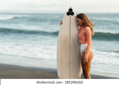 Young attractive surfer woman with white board at sunset on the ocean. Bali Indonesia. Summer time, sports, travel content.