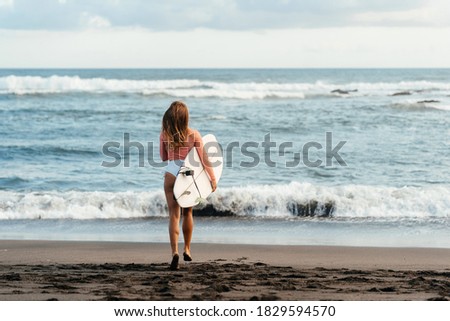 Young attractive surfer woman with white board at sunset on the ocean. Bali Indonesia. Summer time, sports, travel content.