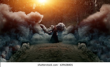 Young attractive Witch in black dress walking on the old bridge in heavy black smoke. Dark magic. Horror fairy tale.