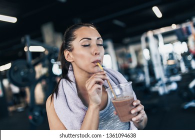 Young Attractive Woman After Successful Workout In Modern Fitness Gym Holding Glass Of Protein Shake And Drinks With Drinking Straw.